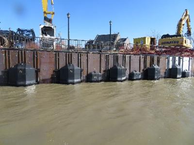 Metal baffle along a riverfront. Construction and excavation equipment on a walkway above the river and behind a railing. The river water is brown.