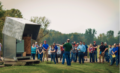 a man in a field gestures at a large piece of equipment before a group of people