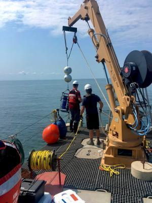 people on a ship operate a crane with scientific equipment being lowered into a lake