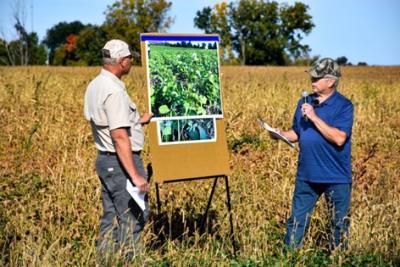 Two people stand in a crop field to give a presentation with a poster board. 