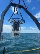 A rosette (sampling equipment) is lifted from the deck of a boat into a clear body of water on a partly cloudy day.